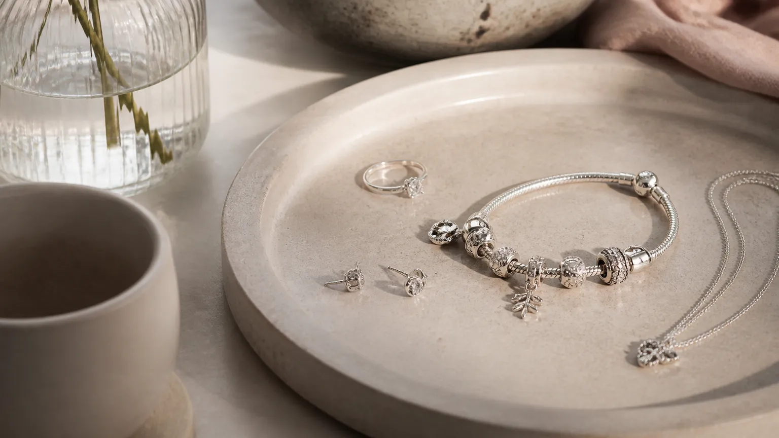 Sølvarmbånd med charms, en ring, to ørestikker og en halskæde med vedhæng ligger på et rundt stenfad ved siden af en keramikkop.
